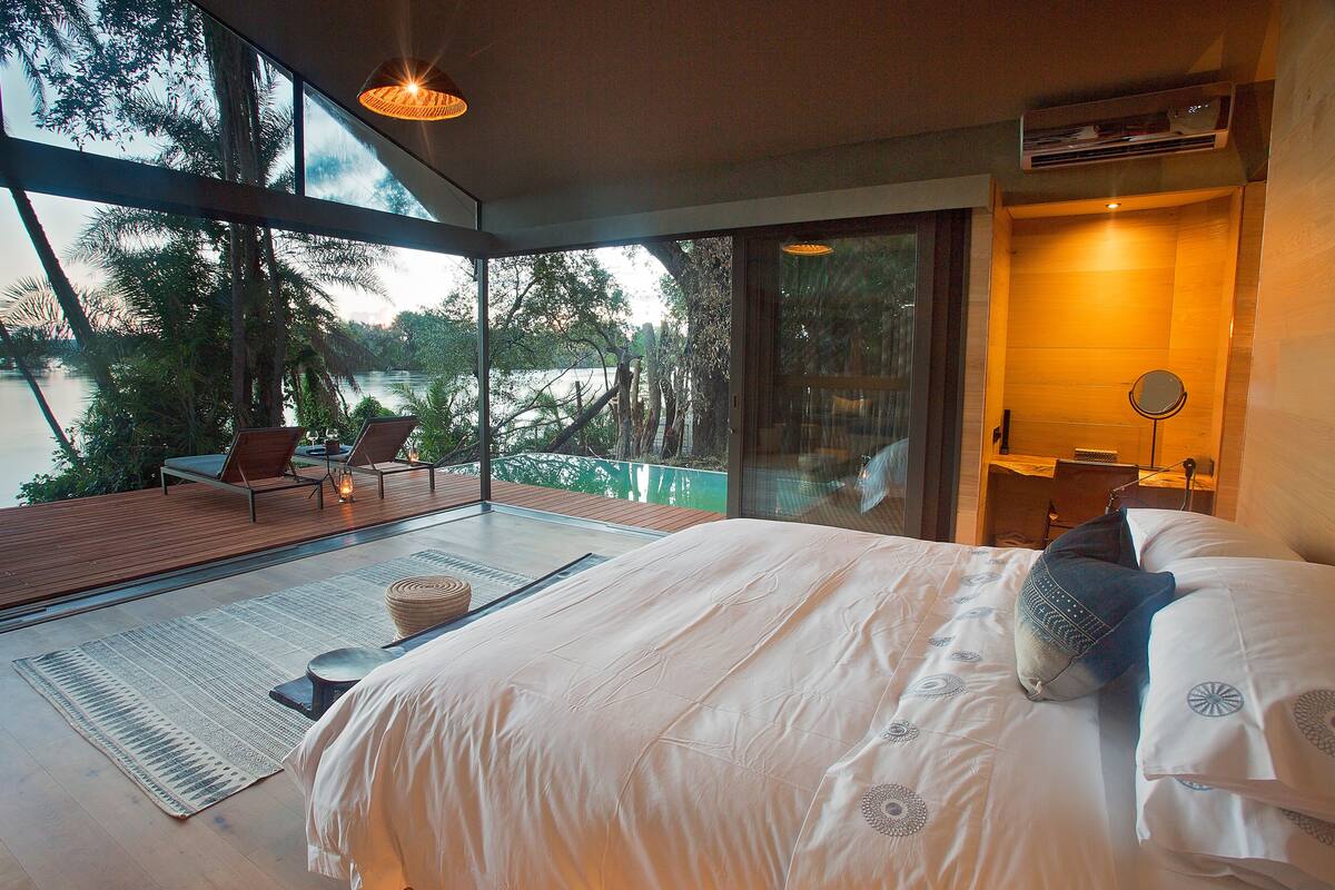 A modern bedroom with a large bed, a view of a lake, and a balcony with chairs.