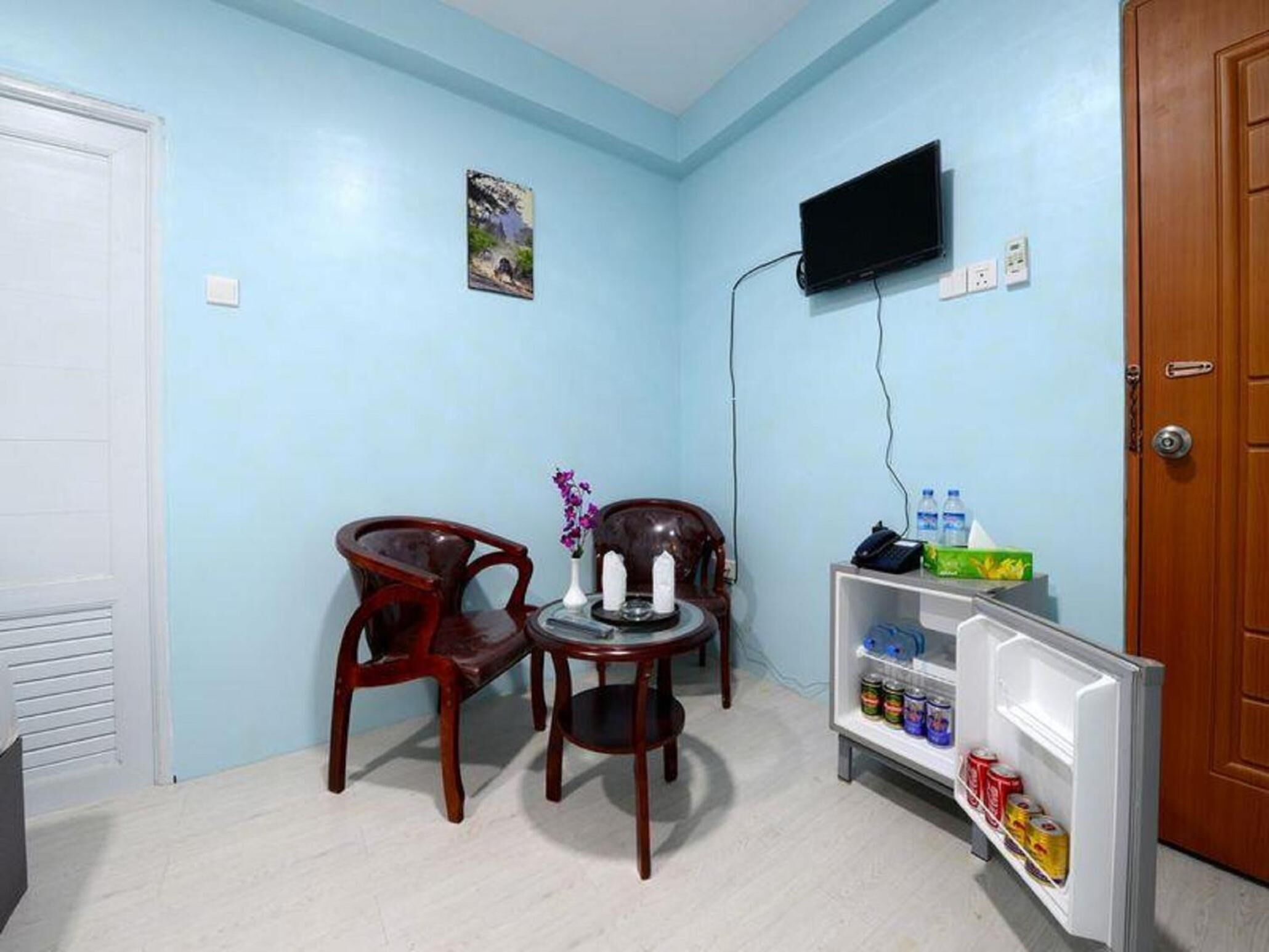 A small room with light blue walls, a wooden door, a television mounted on the wall, two wooden chairs, a small table with a candle and flowers, and a mini-fridge stocked with beverages.