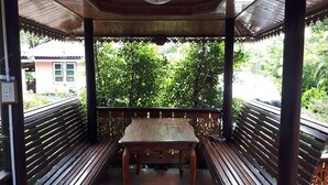 Un espacio de estar al aire libre con techo, bancos de madera y una mesa, rodeado de follaje.