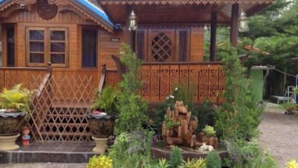 Una casa de madera con porche, cerca de lattice y jardín con diversas plantas y una pequeña formación rocosa.