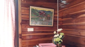 Interior de una cabaña de madera con un cuadro de una casa en la pared, una mesita con un jarrón de flores y un electrodoméstico azul en el suelo.