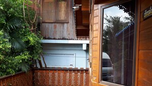 Un balcón de madera con baranda decorativa, una ventana con vistas a árboles y un ramo de flores a la derecha.