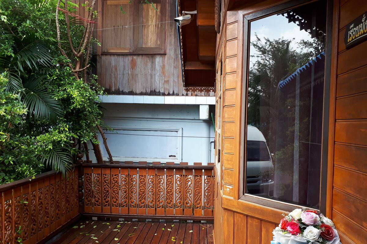 Ein Holzbalkon mit dekorativer Reling, ein Fenster mit Blick auf Bäume und ein Blumenstrauß auf der rechten Seite.