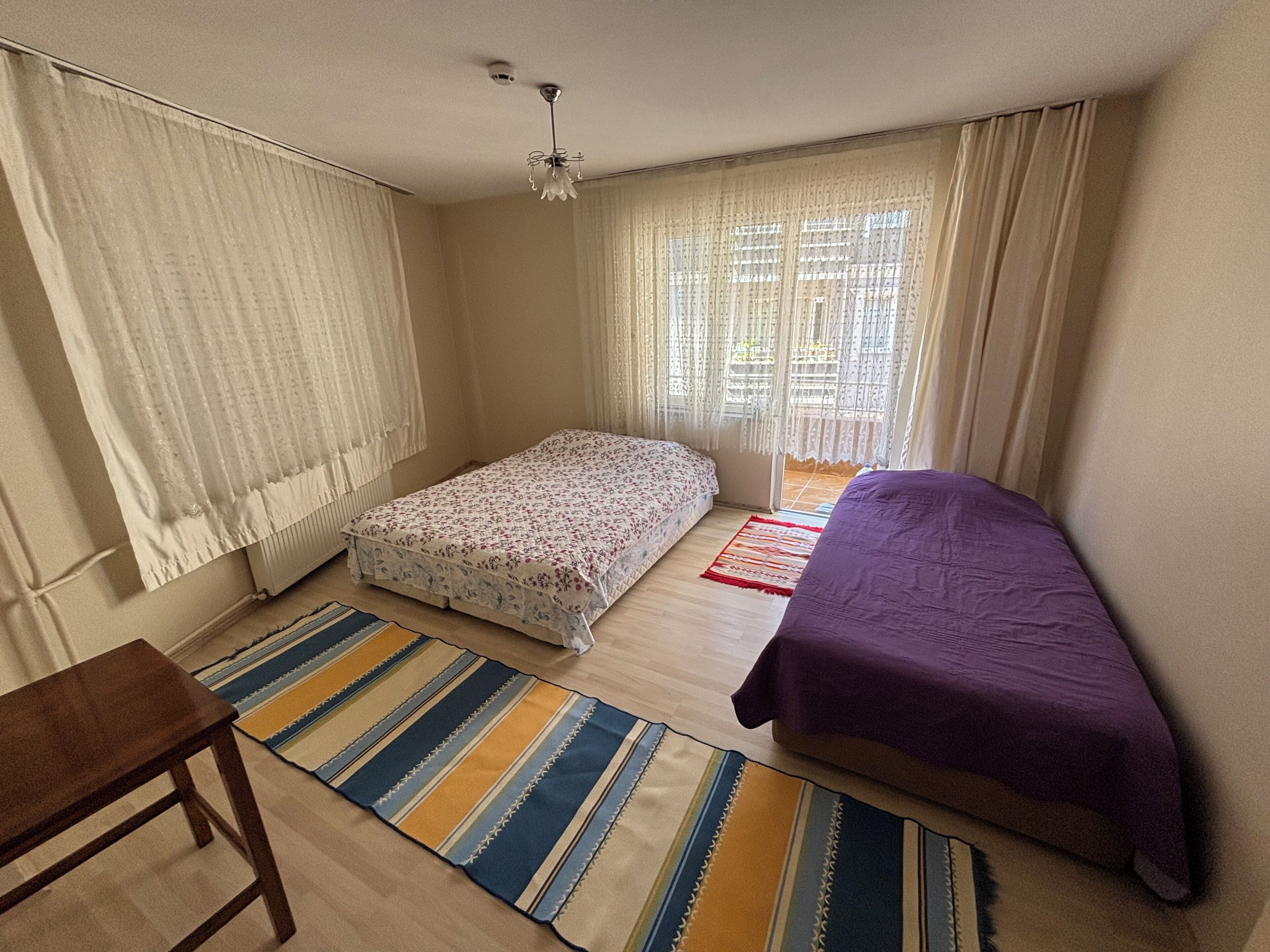 A small, well-lit bedroom with a single bed, a small table, and a window with curtains.