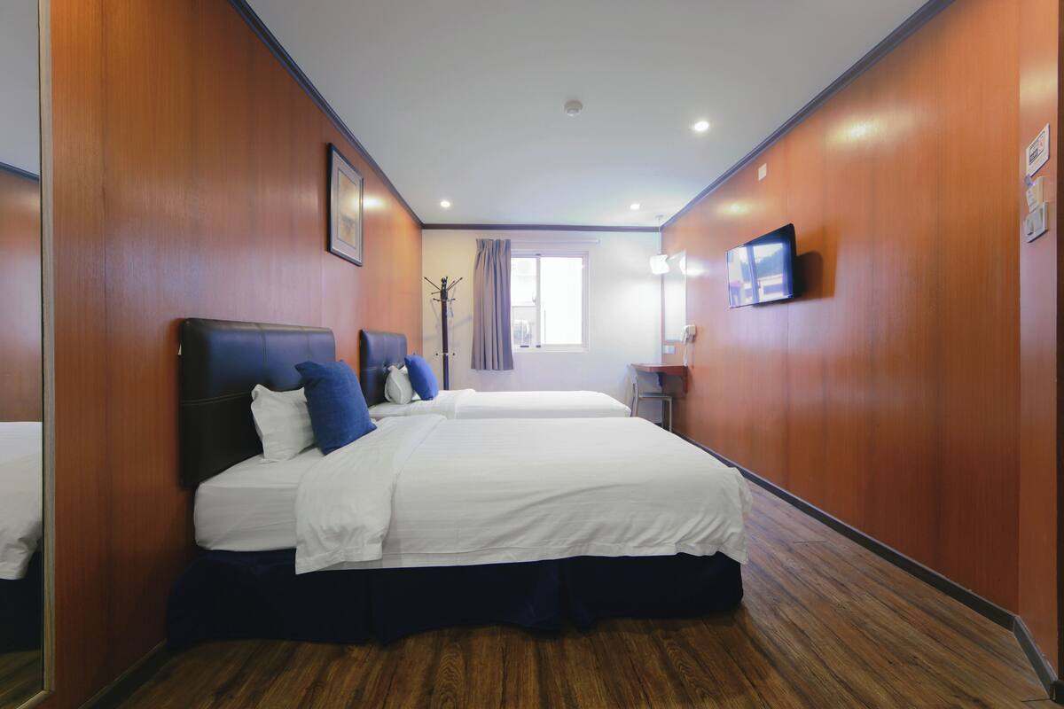 A hotel room with two beds, a flat-screen TV, a desk with a lamp, and a window with curtains.