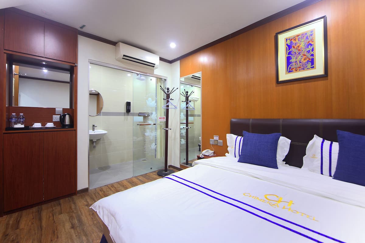 A hotel room with a bed, a wooden wardrobe, a bathroom with a glass shower, and a framed artwork on the wall.