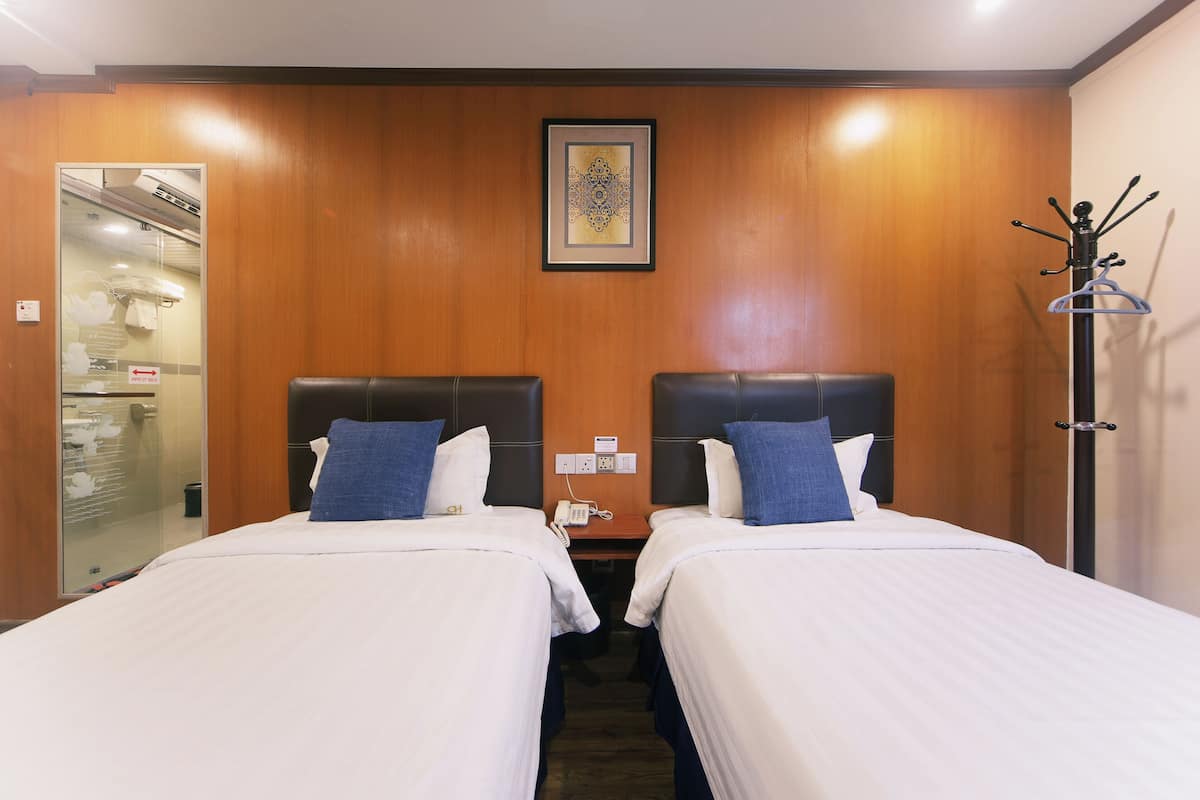 A hotel room with two beds, a wooden headboard, a framed artwork, a coat rack, and a glass door leading to a bathroom.