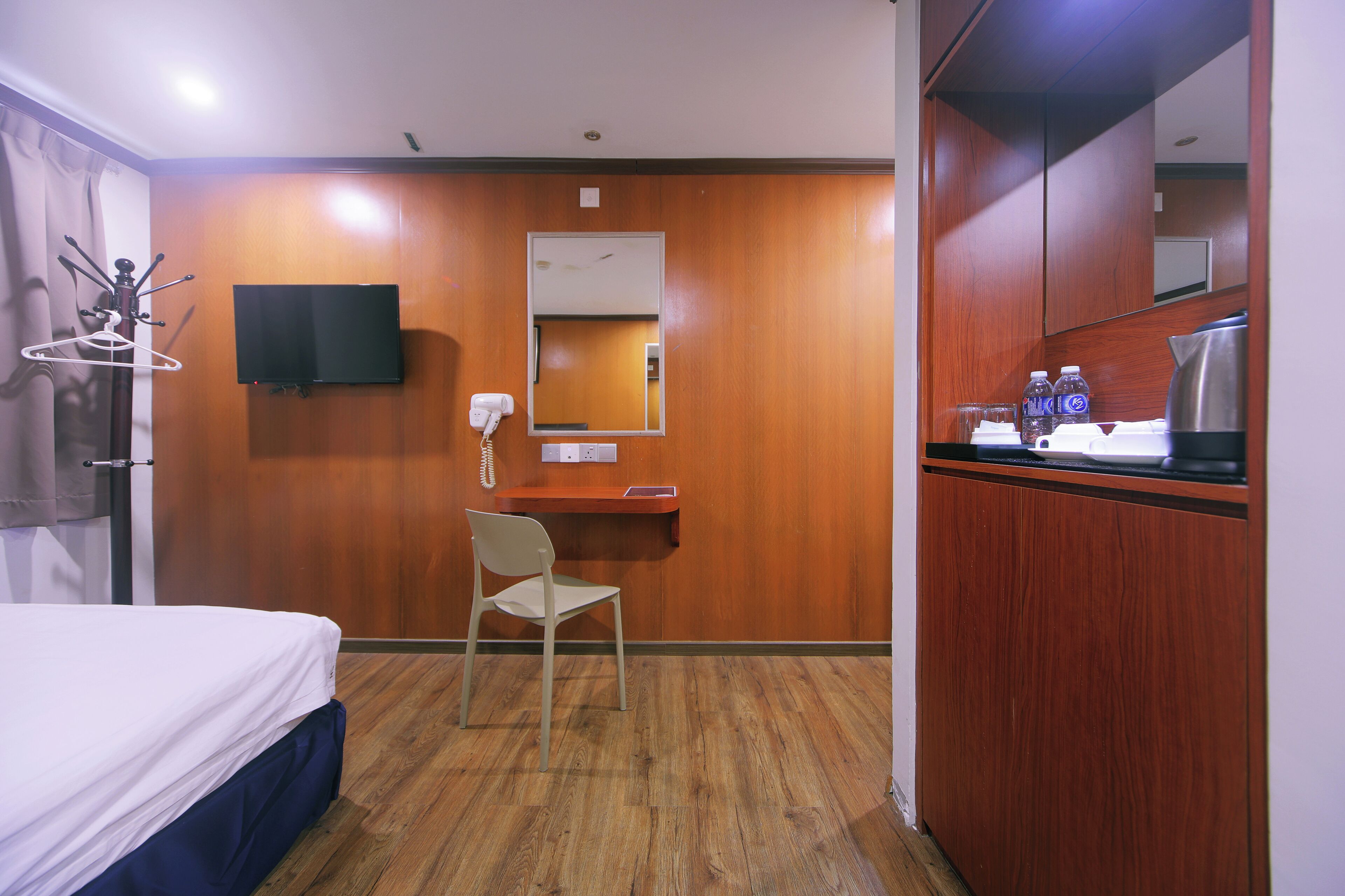 A hotel room with a bed, a desk with a chair, a TV mounted on the wall, a mirror, and a kitchenette area with a coffee maker and bottles.