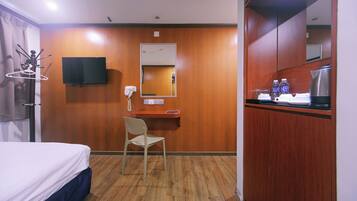A hotel room with a bed, a desk with a chair, a TV mounted on the wall, a mirror, and a kitchenette area with a coffee maker and bottles.