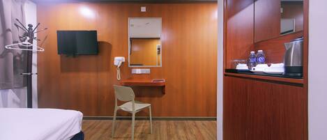 A hotel room with a bed, a desk with a chair, a TV mounted on the wall, a mirror, and a kitchenette area with a coffee maker and bottles.