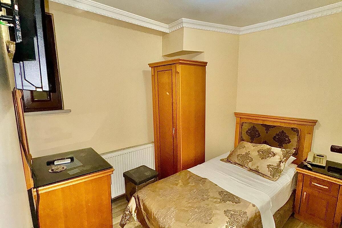 A bedroom with a bed, a wooden wardrobe, a small table, a television mounted on the wall, and a ceiling light.