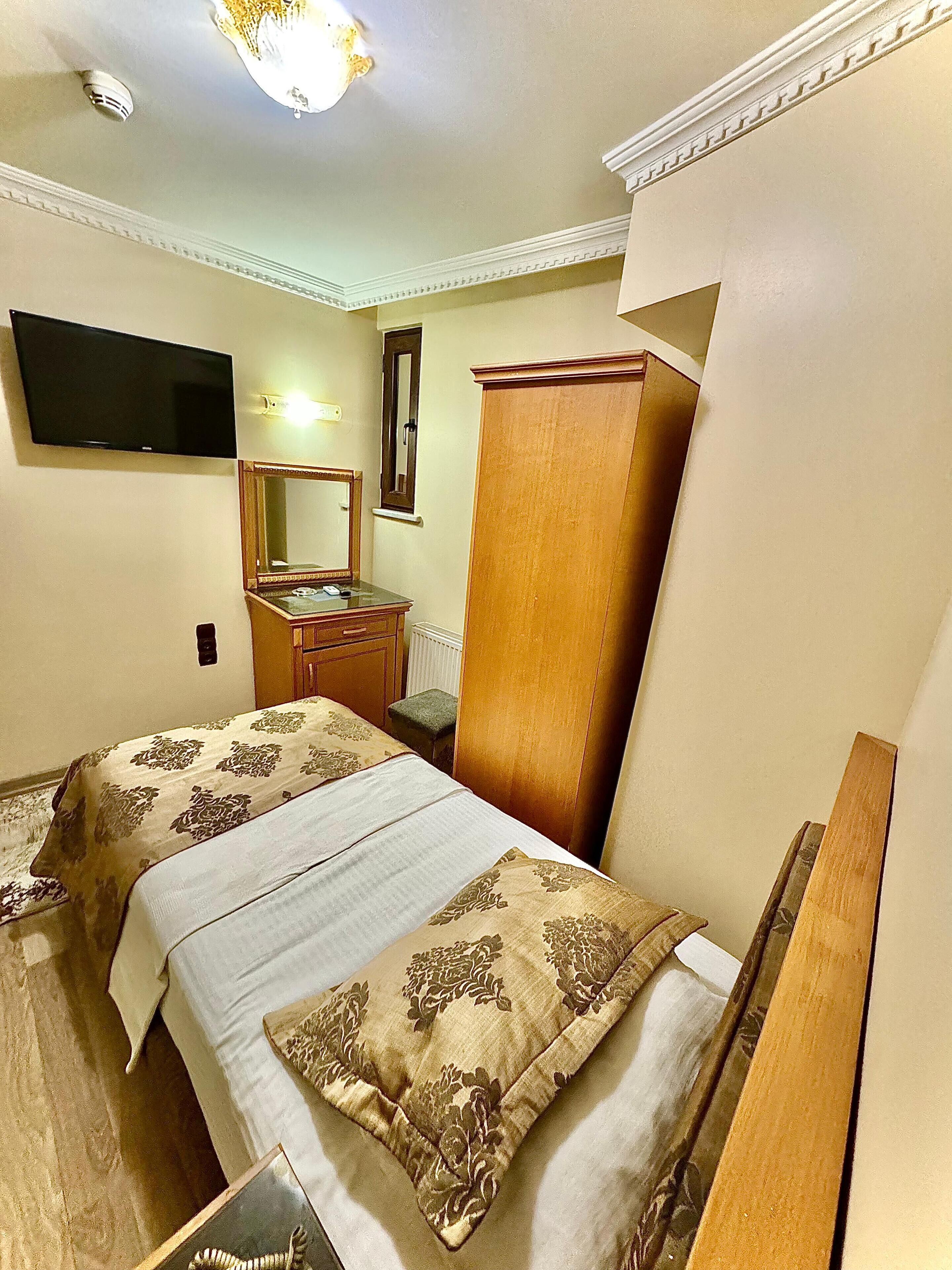 A hotel room with two beds, a wooden wardrobe, a desk with a chair, a flat-screen TV mounted on the wall, and a ceiling light fixture.