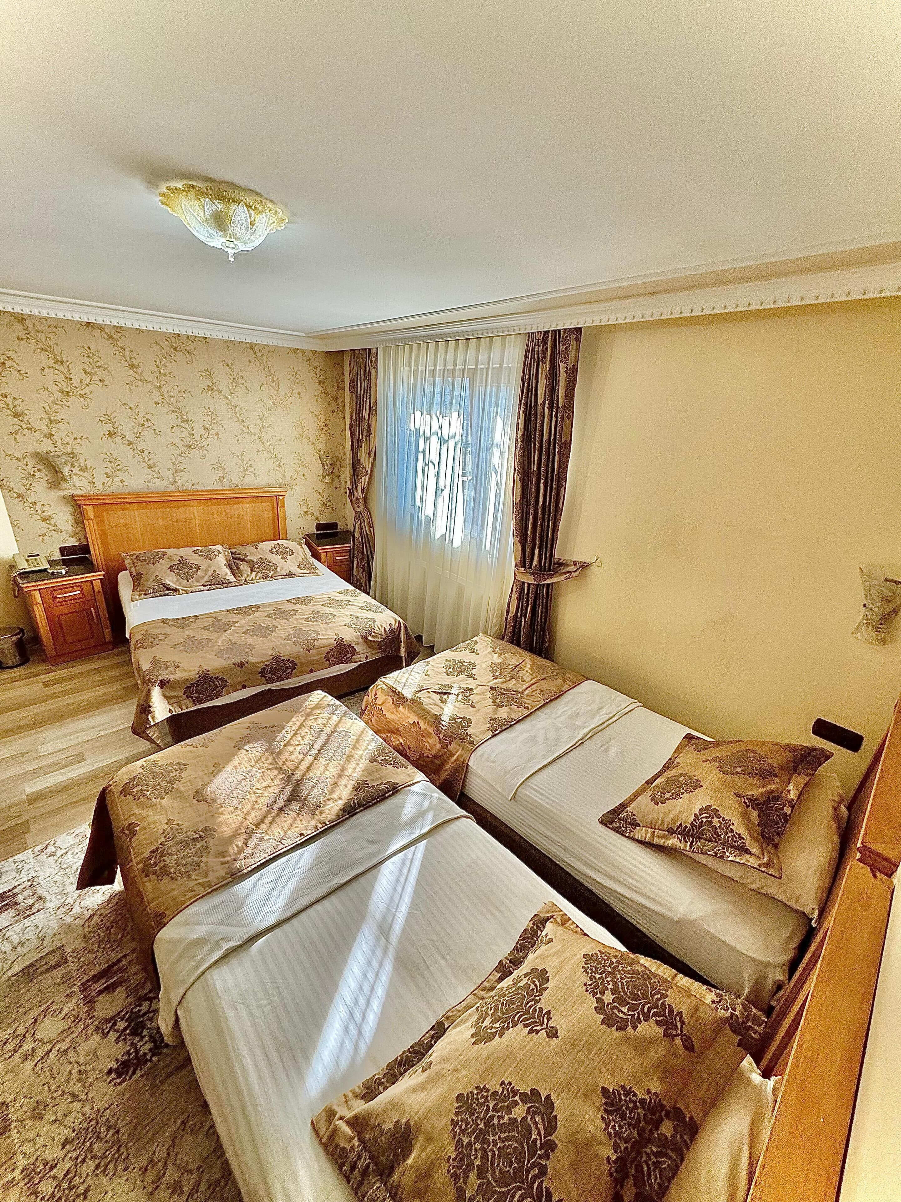 A hotel room with three single beds, a wooden headboard, a bedside table with a lamp, and a window with curtains.