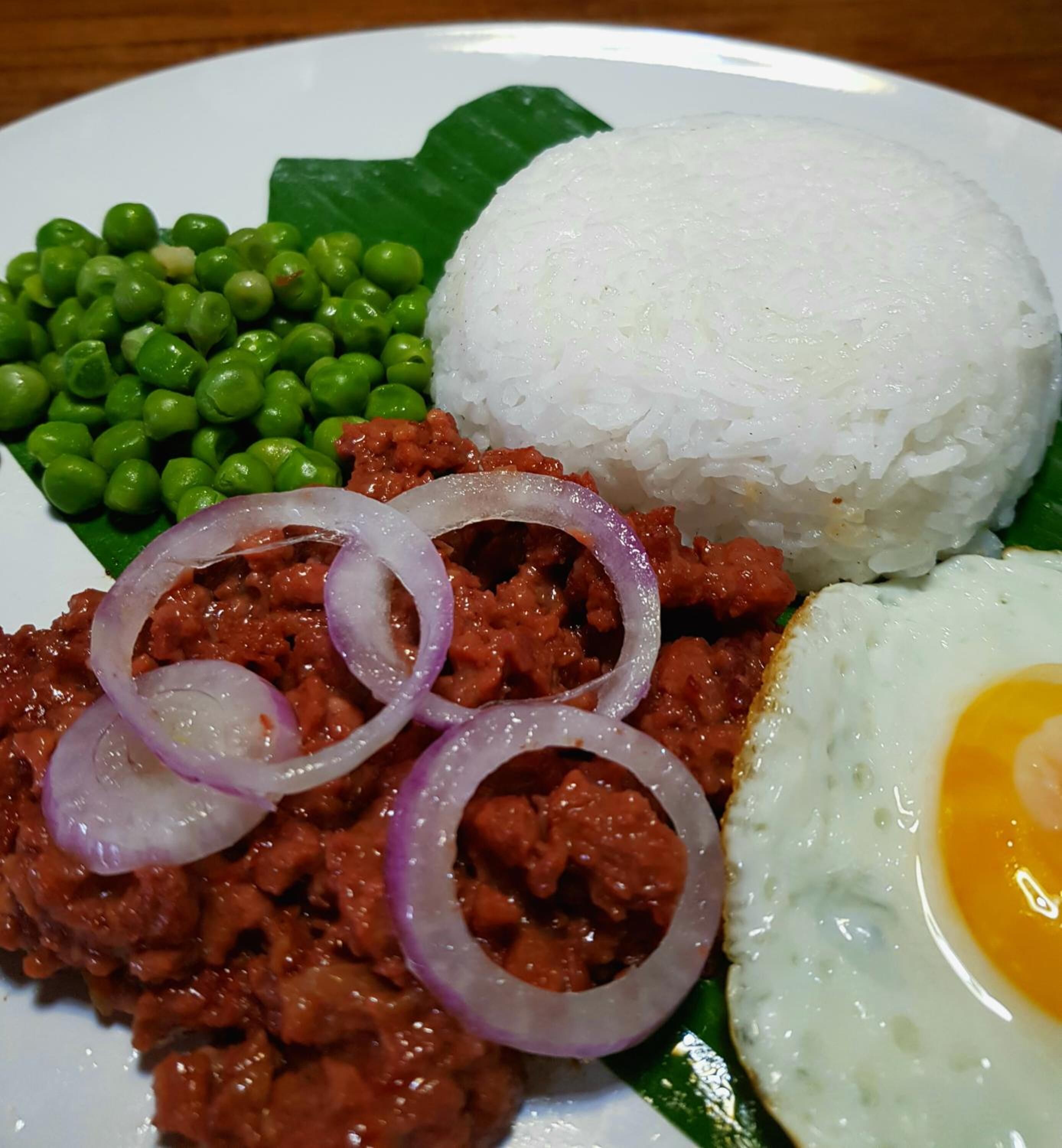 A plate with white rice, green peas, a fried egg, and a meat dish garnished with red onion slices.