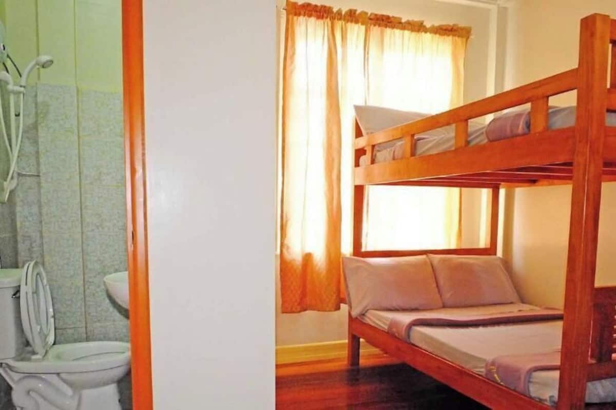 Un baño con ducha, inodoro y lavamanos. Hay una cama litera con colchón y almohadas.