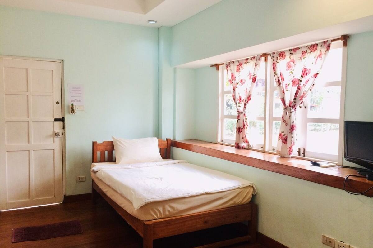 A single bed with a wooden frame, a window with floral curtains, a television on a wooden shelf, and a door with a lock.