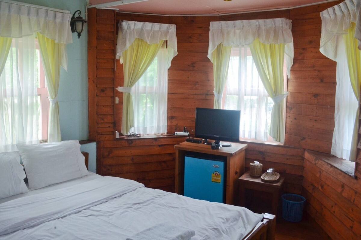 A wooden cabin room with a bed, a television, a small fridge, and a window with curtains.