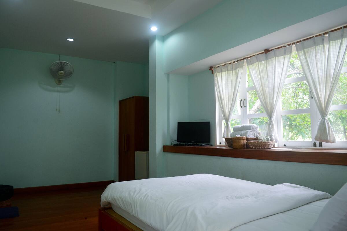 A room with a bed, a television on a shelf, a window with curtains, and a fan on the ceiling.