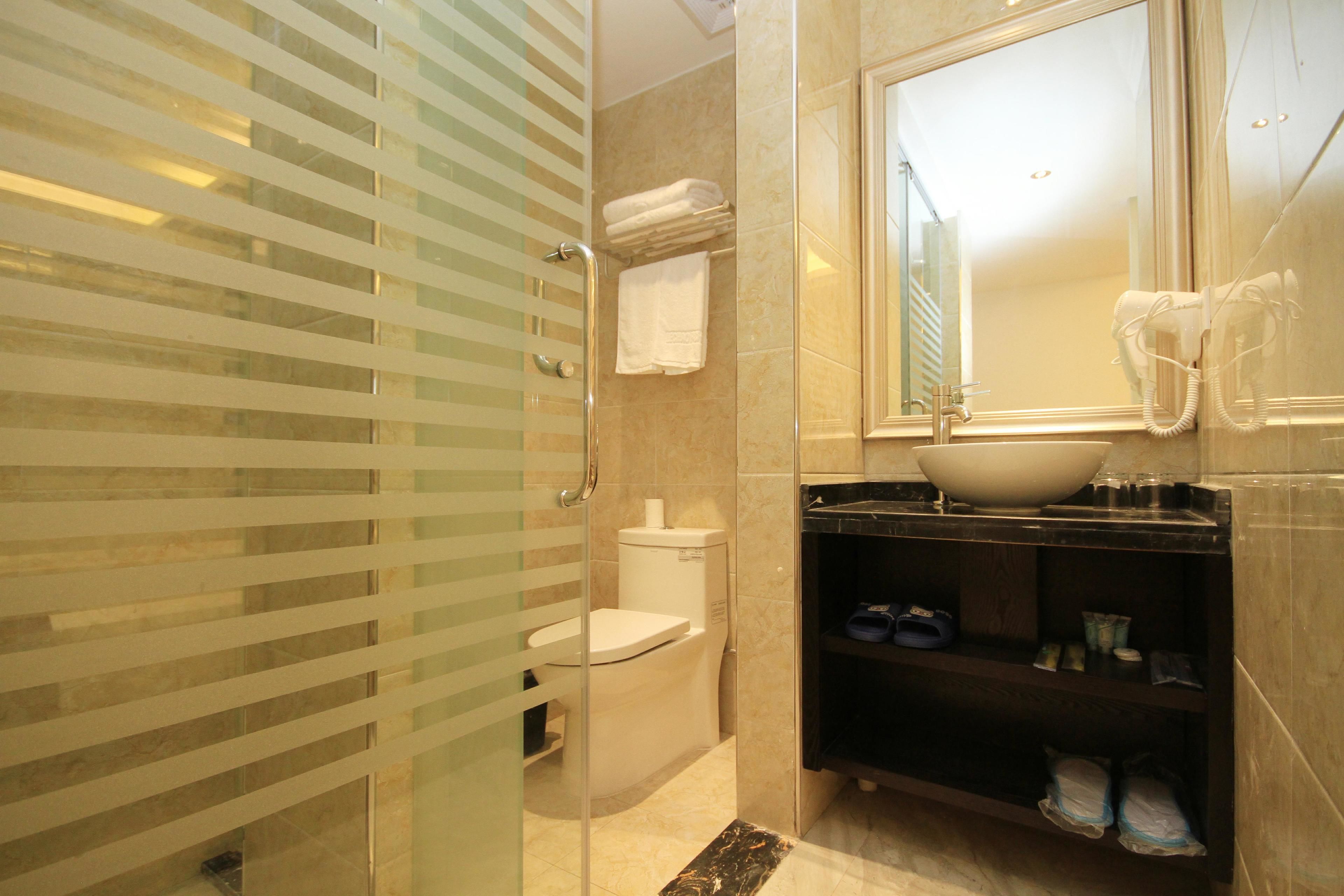 A bathroom with a glass shower enclosure, a toilet, a sink with a large mirror, and a shelf with shoes.