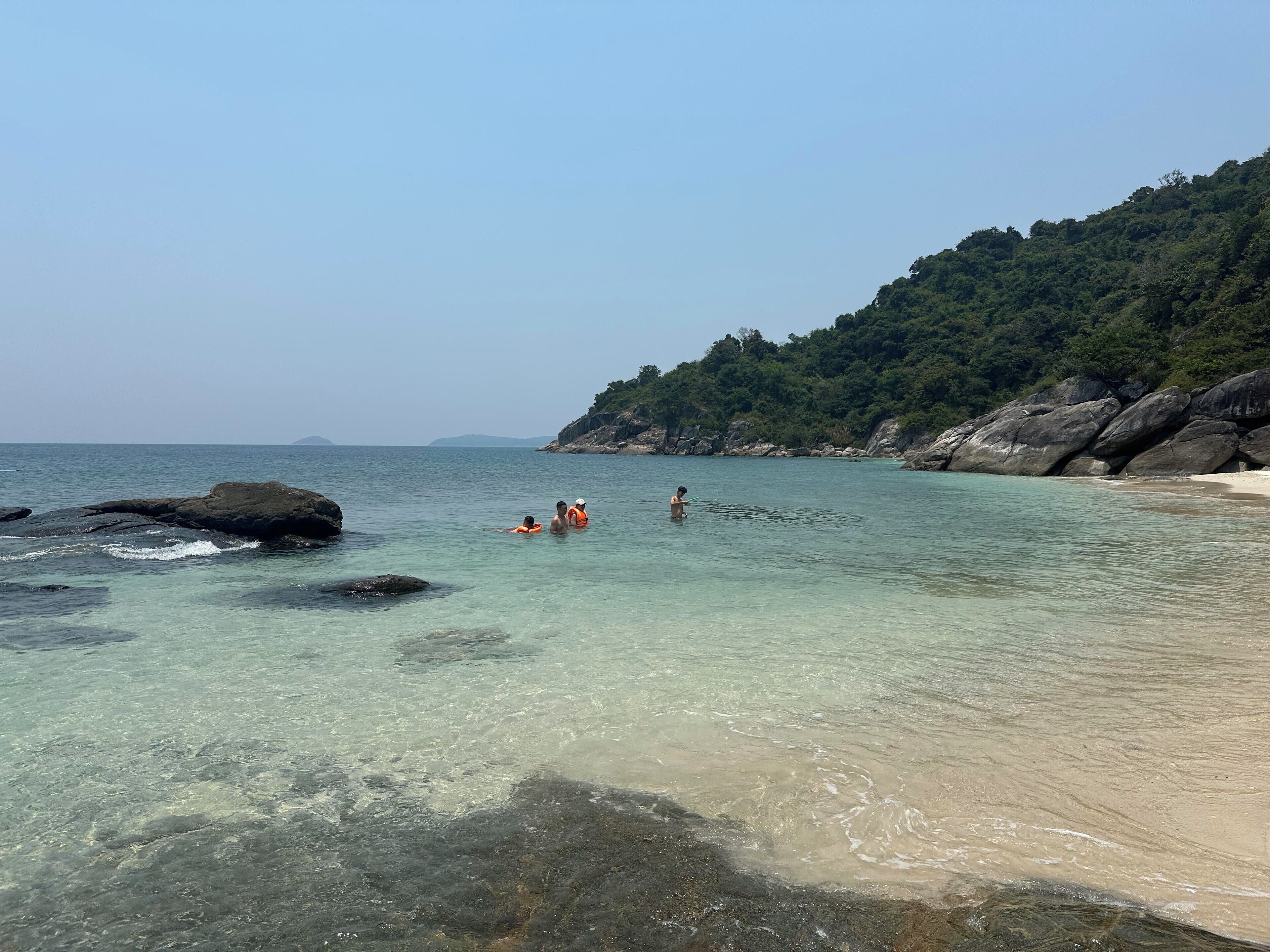 A beach with clear water, rocks, and a forested hill in the background.