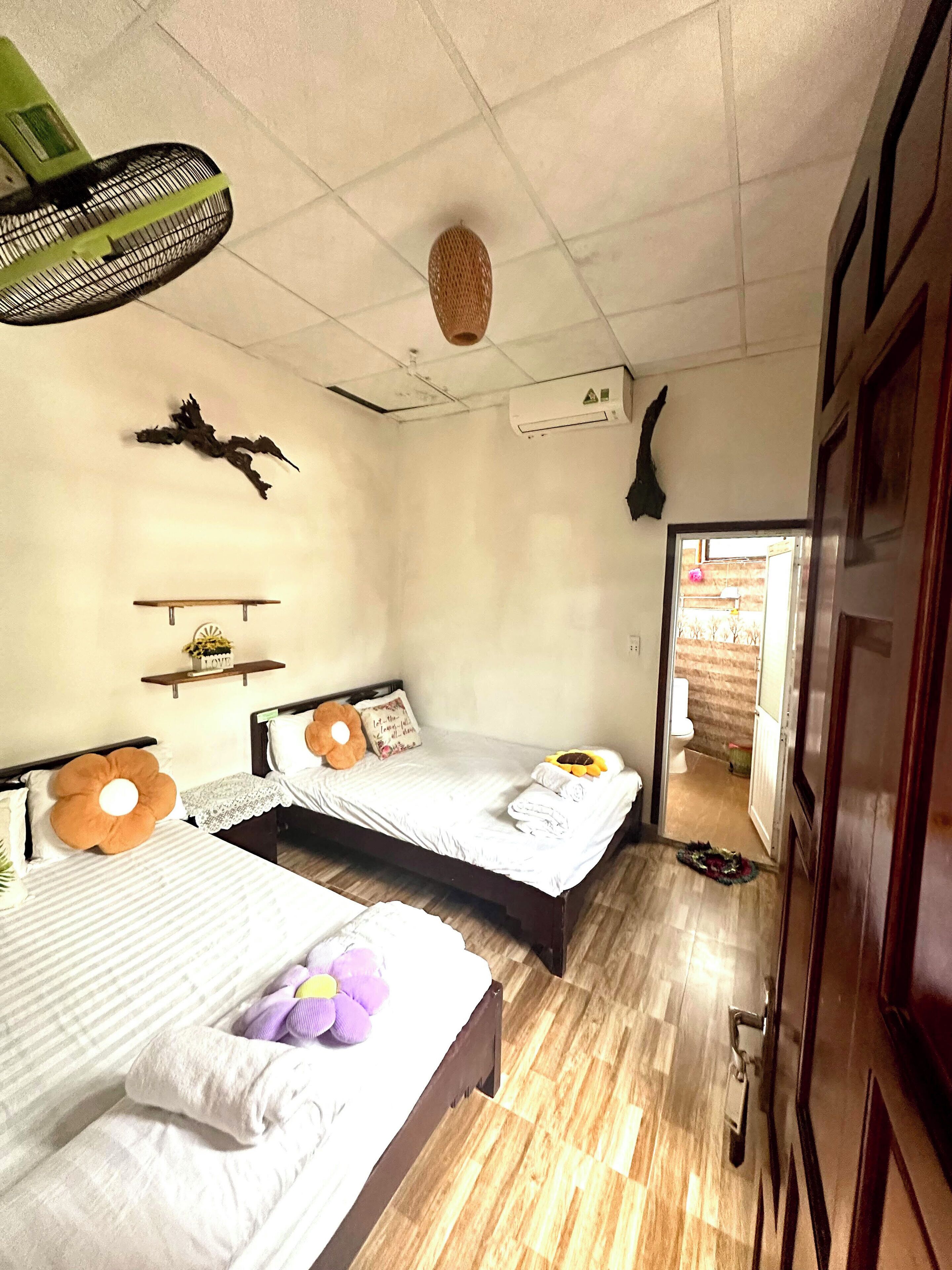 A hotel room with two beds, a ceiling fan, an air conditioning unit, a wall-mounted decoration, and a wooden floor.