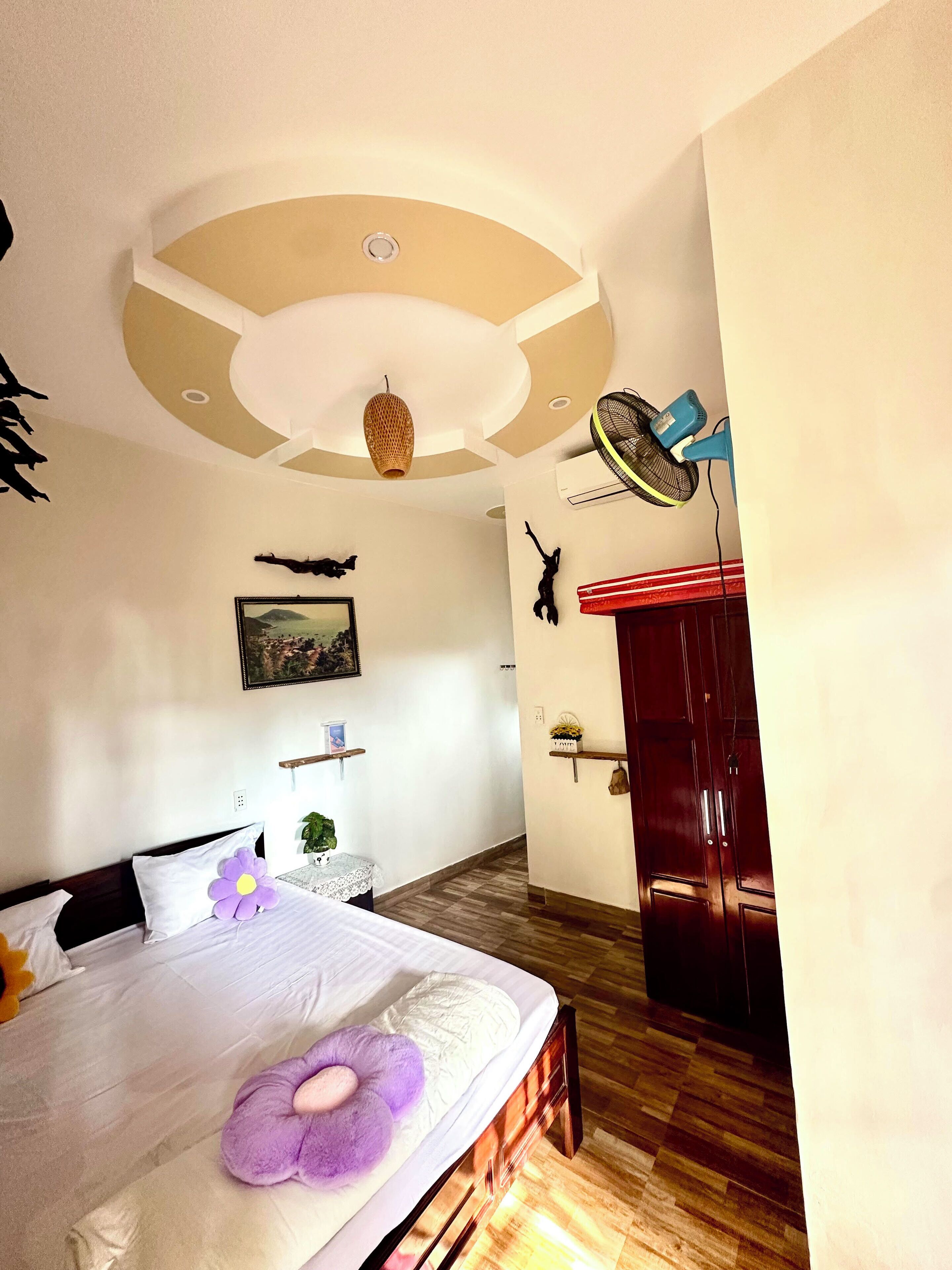 A bedroom with a bed, a ceiling fan, a wooden wardrobe, and a wall-mounted shelf with decorative items.