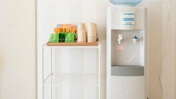 Un dispensador de agua con una jarra grande, un estante blanco con vasos y botellas de agua, y una toma de corriente en la pared.