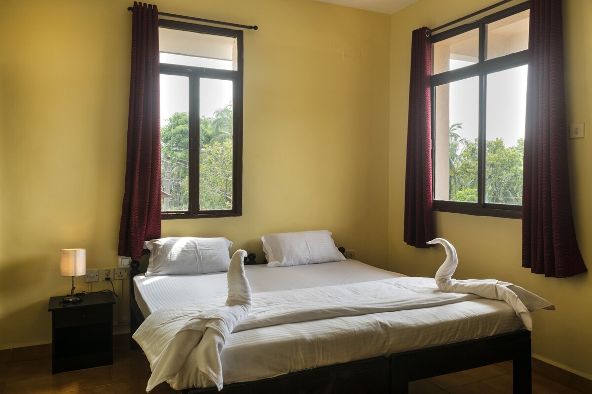 Ein Doppelbett mit weißen Laken und einer zu einem Schwan geformten Handtuch-Arrangement. Ein Nachttisch mit Lampe und ein Fenster mit Vorhängen sind ebenfalls vorhanden.