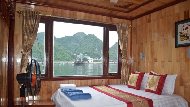 A hotel room with a large bed, a fan, and a view of a body of water and mountains. - Ocean Cruise (Hai Phong)