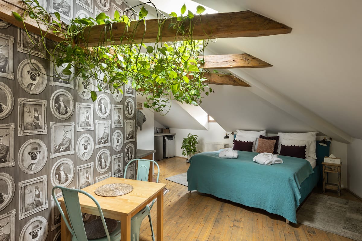 Un dormitorio acogedor en el ático con techo inclinado, cama con cubrecama verde azulado, mesa de comedor con sillas y una pared decorada con cuadros.