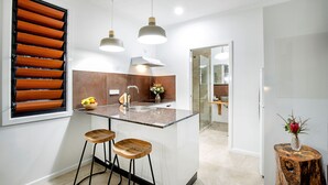 A modern kitchen with a central island, bar stools, pendant lights, and a large window with blinds. - Airlie Beach Eco Cabins - Adults only (Woodwark)