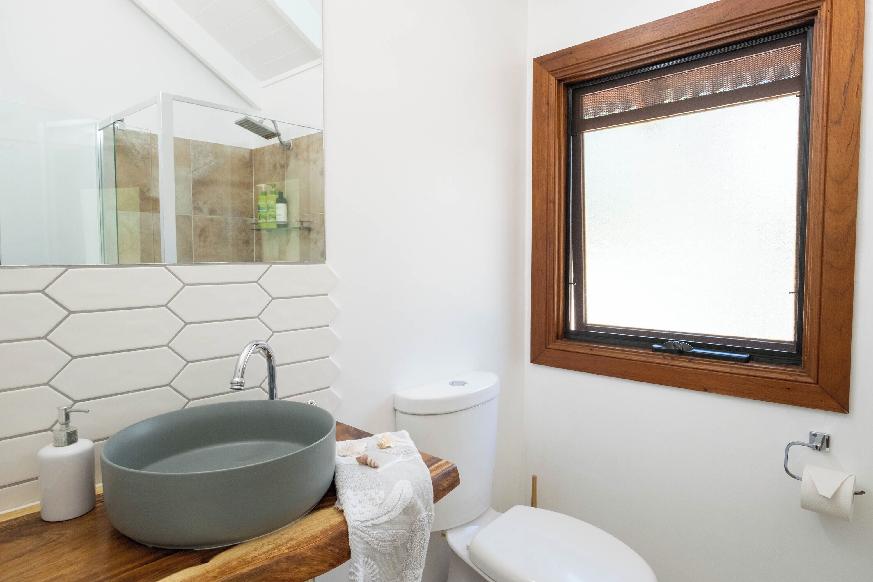 A bathroom with a hexagonal tile backsplash, a round sink, a wooden countertop, a toilet, and a window with a wooden frame.