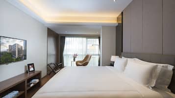 A modern hotel room with a large bed, a desk with books, a chair, and a view of the city through the window.