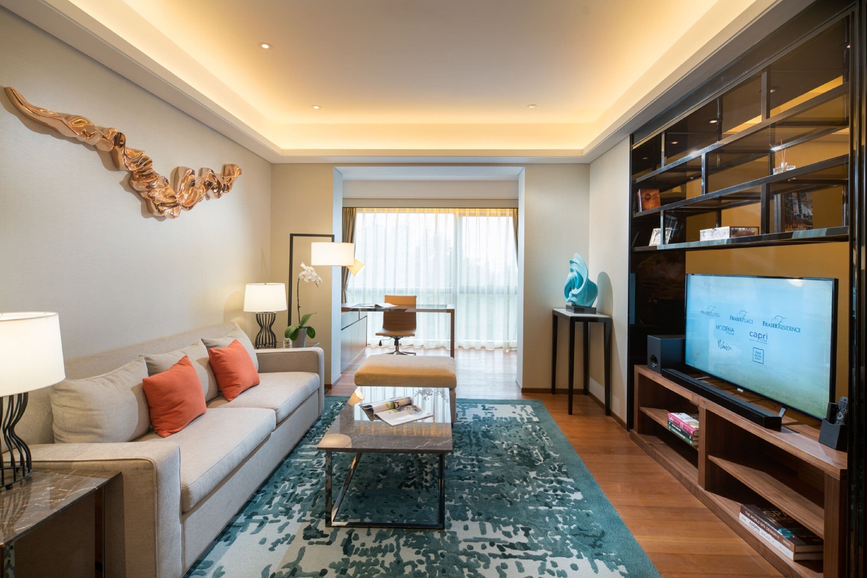 A modern living room with a sofa, coffee table, and TV. There is a bookshelf and a window with curtains.