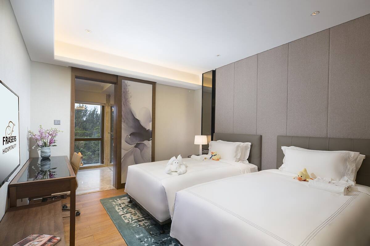 A hotel room with two beds, a desk, a lamp, and a view of the outdoors.