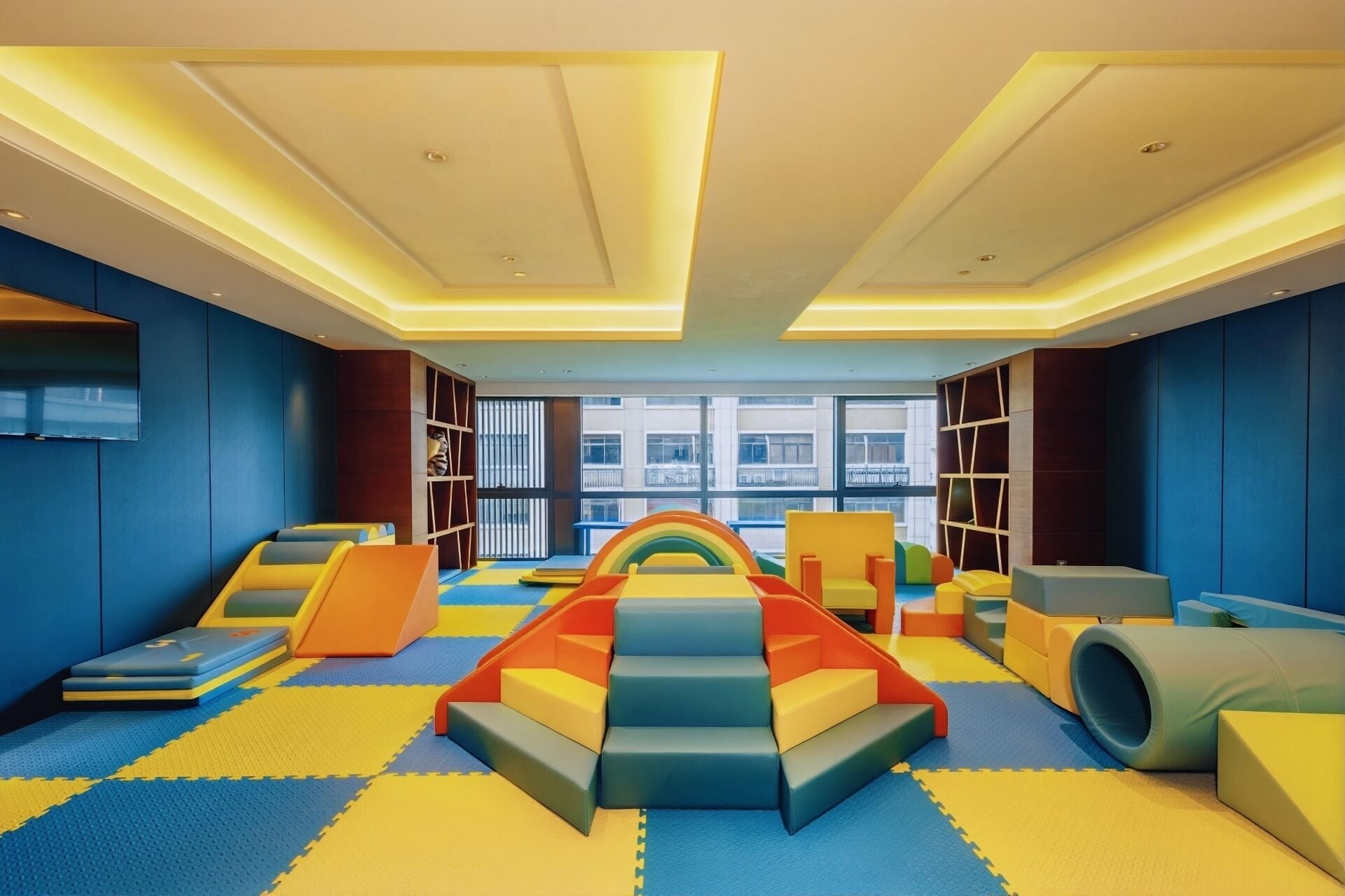 Zona infantil con estructuras de juego coloridas, suelo acolchado y grandes ventanales con vistas al exterior.