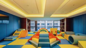 Zona infantil con estructuras de juego coloridas, suelo acolchado y grandes ventanales con vistas al exterior.