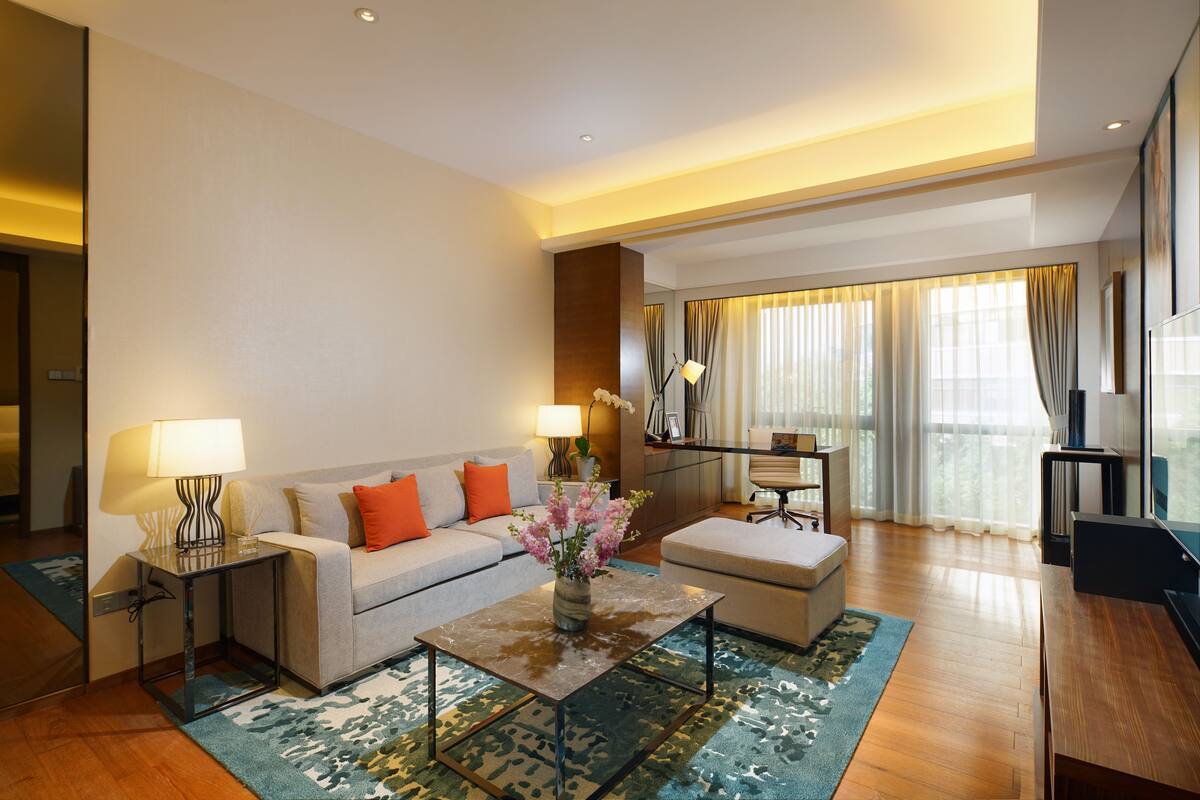 A modern living room with a sofa, coffee table, and lamps. There is a large window with curtains and a view of greenery outside.