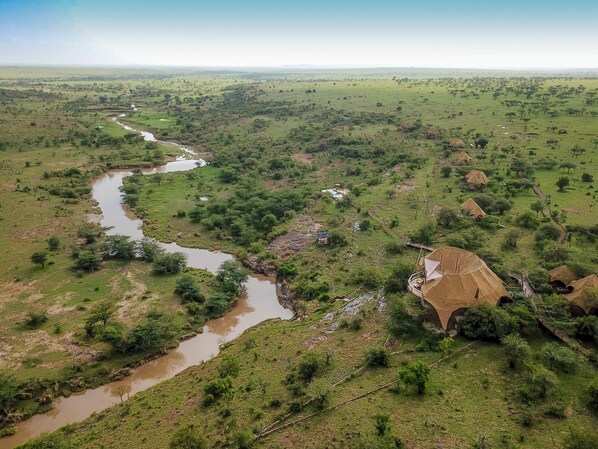 An aerial view of a rural landscape with a river, scattered trees, and traditional huts. - Amani Mara Camp (Maasai Mara)