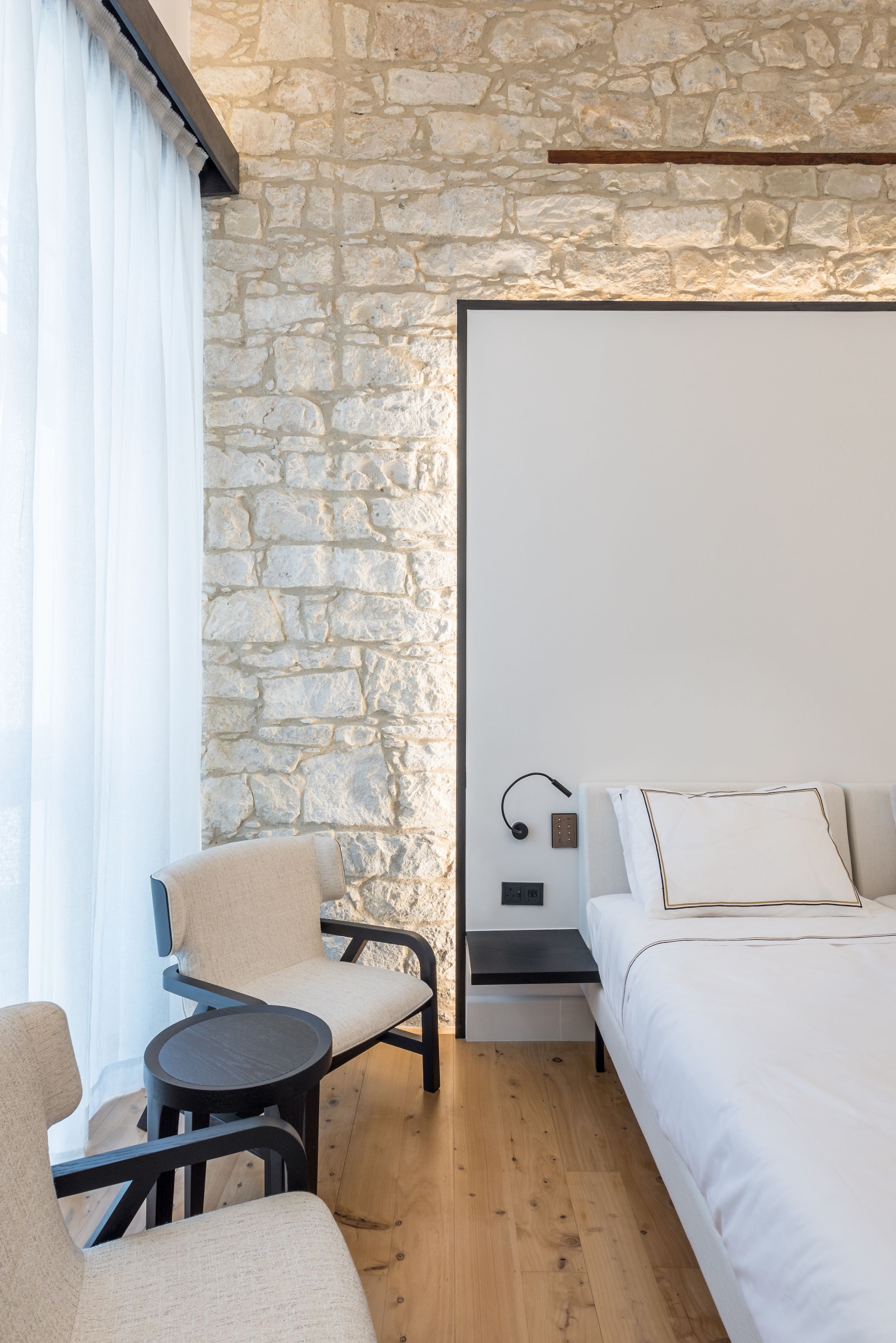 A hotel room with a stone wall, a bed, a chair, a small table, and a bedside table with a lamp.