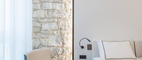A hotel room with a stone wall, a bed, a chair, a small table, and a bedside table with a lamp.
