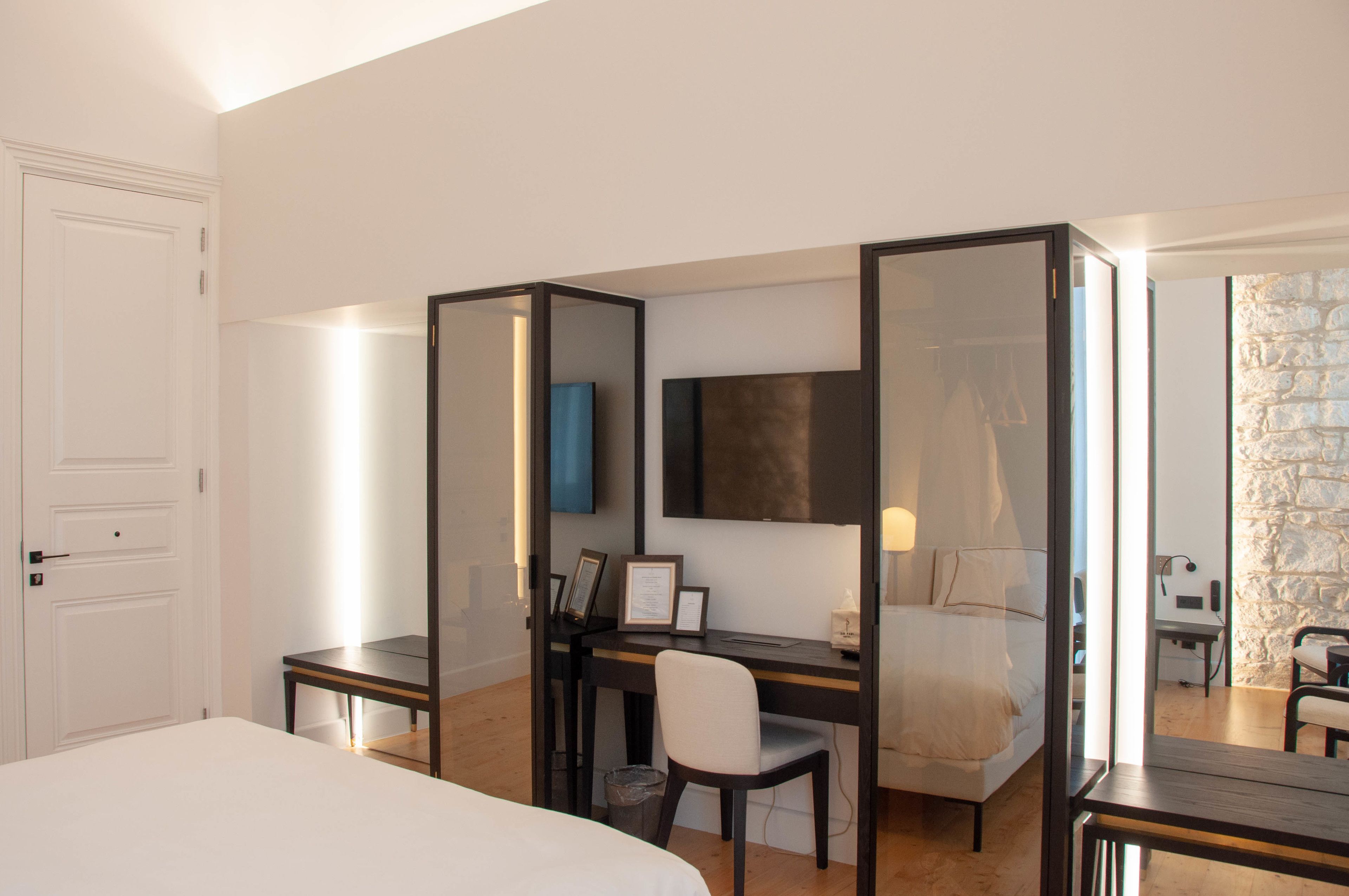 A hotel room with a bed, a desk with a chair, a TV mounted on the wall, and a stone wall.