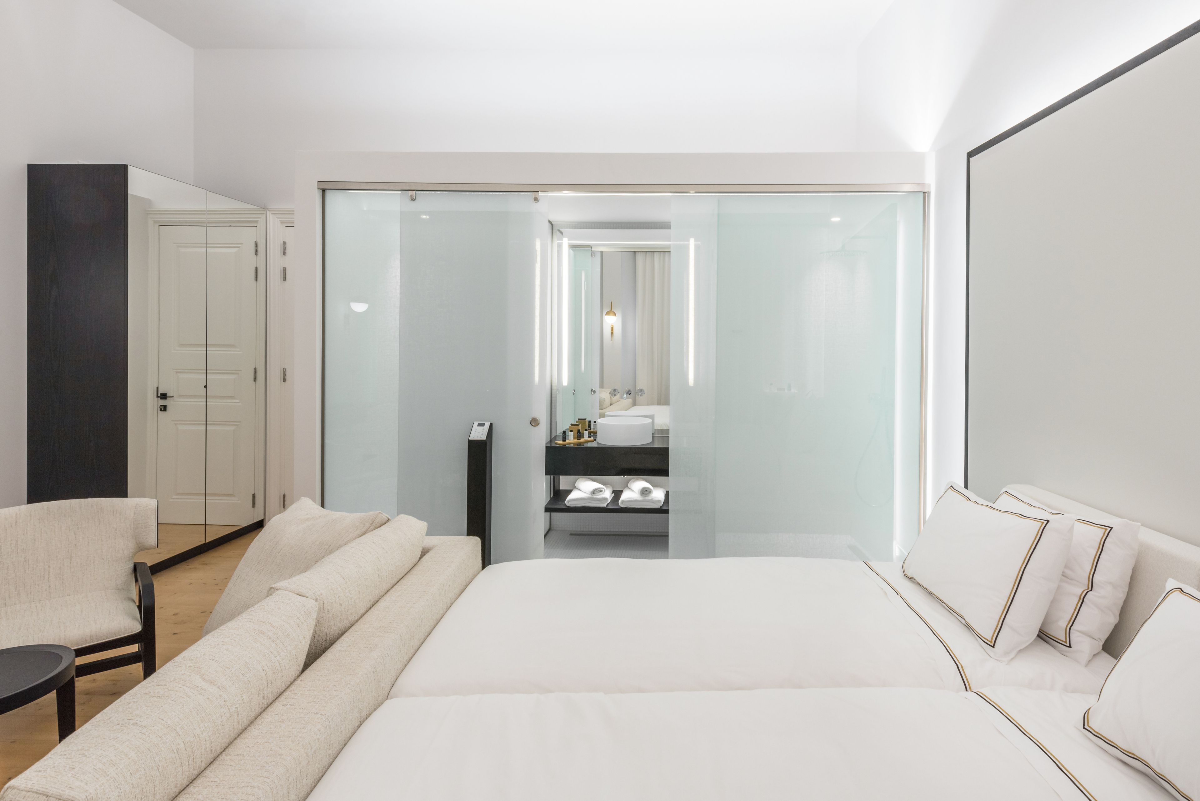 A modern hotel room with a large bed, a sofa, a chair, a glass-enclosed bathroom, and a wardrobe.