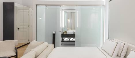 A modern hotel room with a large bed, a sofa, a chair, a glass-enclosed bathroom, and a wardrobe.