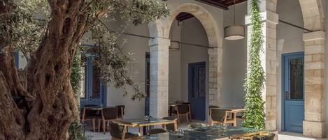 An outdoor patio with stone arches, wooden tables, and chairs, surrounded by greenery and a large tree.