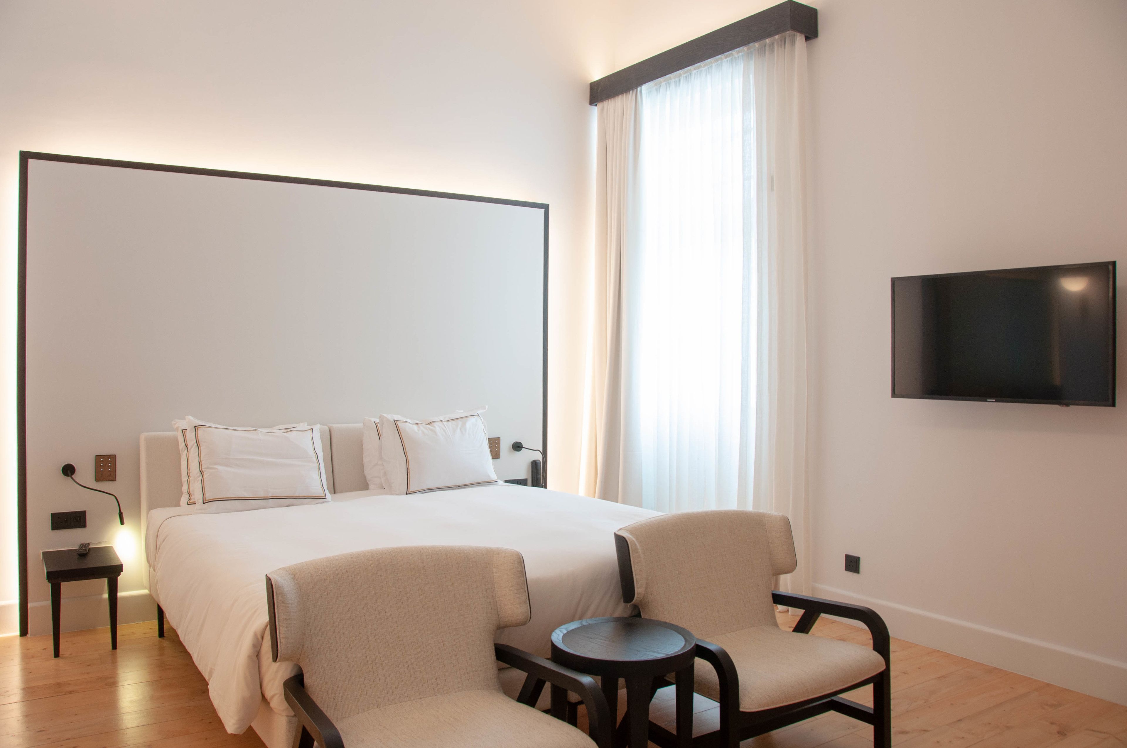 Habitación de hotel con una cama grande, dos sillones, una mesita, televisor montado en la pared y una ventana con cortinas.