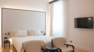 Habitación de hotel con una cama grande, dos sillones, una mesita, televisor montado en la pared y una ventana con cortinas.