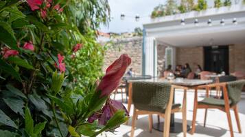 Un patio al aire libre con sillas y mesas de madera, rodeado de abundante follaje y plantas con flores.