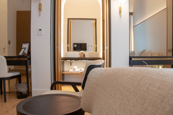 A hotel room with a bed, a round table, a chair, a vanity with a mirror, and a small shelf with towels. - S Paul Hotel (Limassol)