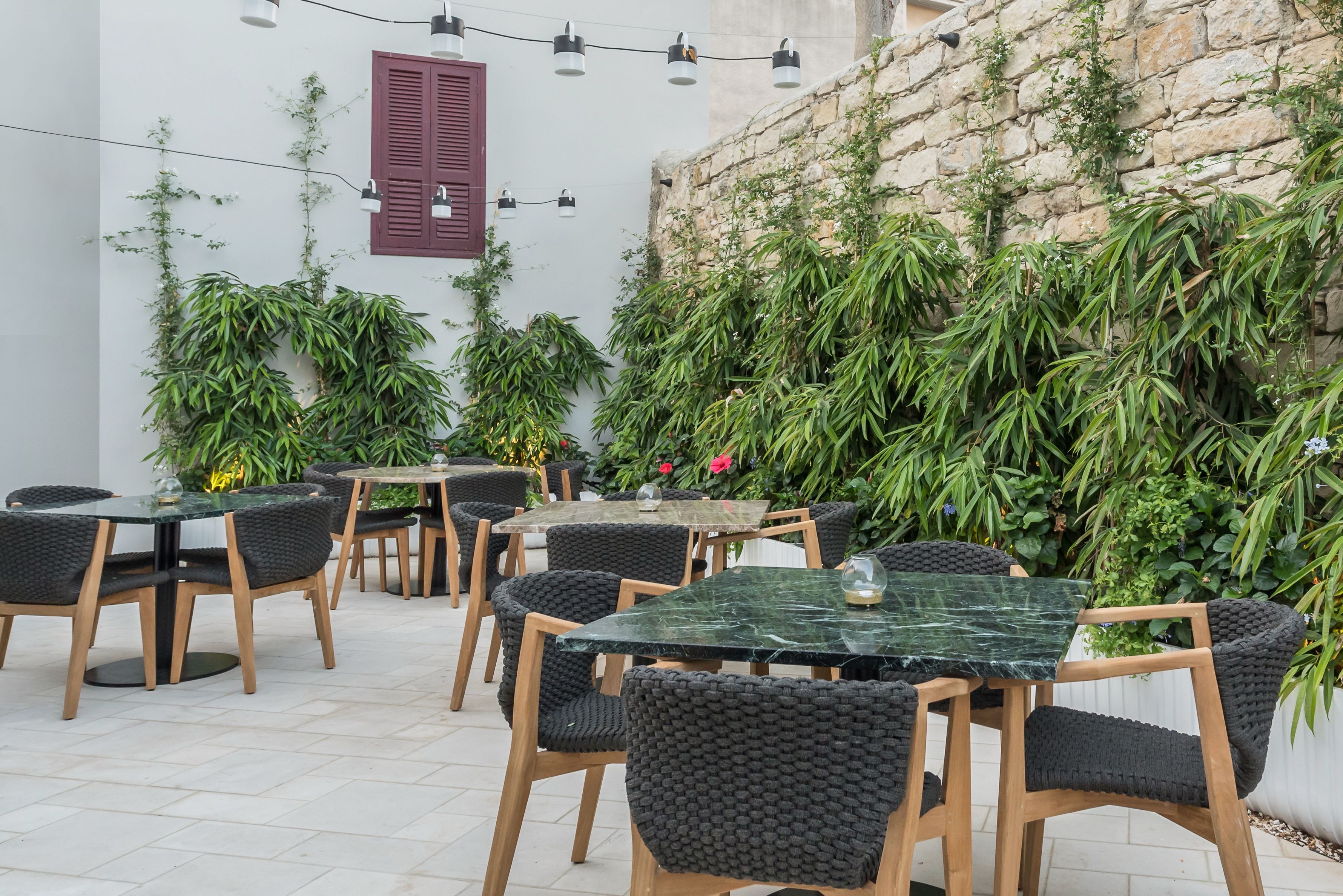 An outdoor seating area with tables and chairs, surrounded by lush greenery and a stone wall.